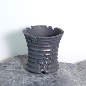 Peut inclure: Un vase en céramique gris foncé avec un design en spirale texturé. Le vase a une forme cylindrique avec un bord légèrement irrégulier. La surface a un aspect rugueux, presque vieilli, ajoutant à son esthétique unique. Le vase est posé sur une surface en pierre grise.