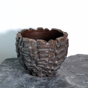 May include: A textured, bronze-colored ceramic planter with a rough, brick-like exterior. The planter has a dark brown interior and an uneven rim. It sits on a dark gray stone surface against a white background.