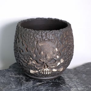 Peut inclure: Un vase en céramique gris foncé avec un extérieur texturé. Le vase présente un motif de crâne sculpté avec des flammes et des cornes. L'intérieur est lisse et sombre. Le vase est posé sur une surface en pierre grise foncée.