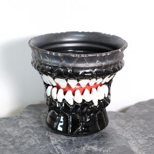Peut inclure: Un vase en céramique noir avec un design unique en forme de bouche ouverte. Le vase présente des dents blanches, un intérieur rouge et un extérieur texturé. Le bord est gris foncé et le vase est posé sur une surface grise.