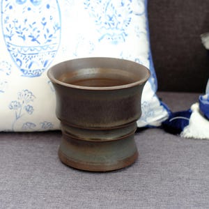Peut inclure: Un vase en céramique marron avec un dessus évasé et un corps cylindrique strié. Le vase a une couleur sombre et terreuse avec de subtiles variations. Le design est simple et élégant, adapté pour exposer des fleurs ou comme pièce décorative.