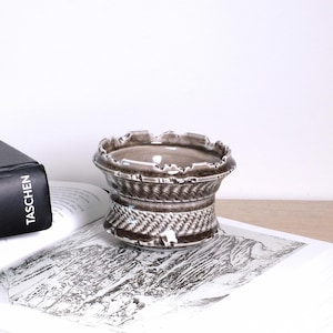 以下が含まれることがあります： 茶色とグレーの色合いの、バスケット織りのテクスチャデザインが施された、小さな装飾用セラミックボウル。 ボウルにはスカラップの縁があり、本の上に置かれています。 「TASCHEN」という単語が見えます。 ボウルは、小さなアイテムを保持するために使用される可能性があります。