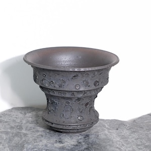 Puede incluir: Un jarrón de cerámica gris oscuro, hecho a mano, con un borde ancho y acampanado y una superficie texturizada y picada. El jarrón tiene un cuerpo cilíndrico con una serie de bandas decorativas y un interior oscuro. La pieza se exhibe sobre una superficie de piedra gris.