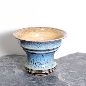 May include: A handcrafted ceramic bowl with a wide, shallow rim and a flared base. The bowl features a gradient of colors, with a tan interior, a blue-toned middle section, and dark brown accents. The bowl is likely used for decorative purposes.