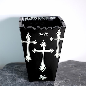 Peut inclure: Pot de fleurs noir avec des croix blanches et les textes "PLANTS NEVER DIE" et "SAVE". Le pot a un bord supérieur dentelé unique et une finition brillante. Conçu pour les plantes.