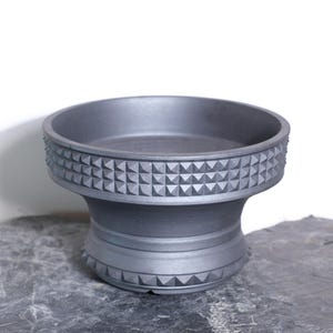 May include: A dark gray ceramic bowl with a wide, shallow basin and a pedestal base. The rim and base feature a repeating geometric pattern of small, raised triangles. The bowl is approximately 6 inches in diameter and 4 inches tall.