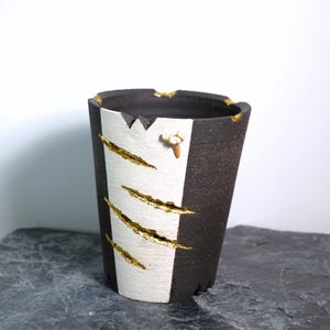 Peut inclure: Un vase en céramique fait à la main avec un design à rayures noires et blanches. Des accents de feuilles d'or ornent la bande blanche, avec un petit élément décoratif sur le côté. Le vase a une surface texturée et une esthétique artistique unique.