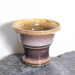 May include: A handcrafted ceramic vase with a flared top and a pedestal base. The vase features a gradient of colors, transitioning from a golden-brown rim to a deep purple-brown base. The interior is a glossy brown.