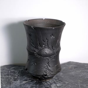 Peut inclure: Un vase en céramique noire avec une surface texturée et un motif d'œil. Le vase a une large ouverture et une forme légèrement évasée. L'œil est au centre du vase, avec des fissures rayonnantes.