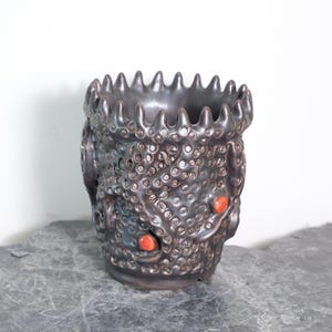 Pode incluir: Um vaso de cerâmica cinza escuro com uma superfície texturizada e uma borda irregular. O vaso apresenta um padrão de pequenos círculos e é acentuado com dois detalhes circulares vermelhos. O vaso é exibido em uma superfície cinza.
