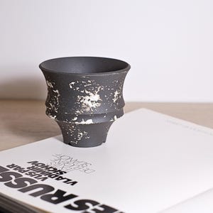 Puede incluir: Un jarrón de cerámica negro con un patrón moteado blanco. El jarrón tiene un borde acampanado y un cuerpo cilíndrico estriado. Se encuentra sobre un libro blanco con texto negro. El jarrón mide unos 10 cm de alto.