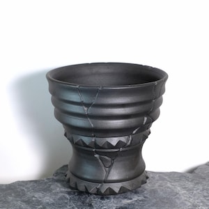 May include: A dark gray ceramic vessel with a unique, textured design. The vessel features a series of horizontal ridges and a base with triangular patterns. Visible cracks add to the piece's character. The pottery is displayed on a gray stone surface.