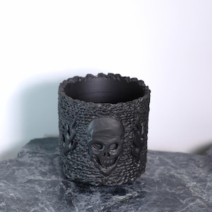 Peut inclure: Un récipient cylindrique noir avec une surface texturée. Le récipient présente un motif de crâne et de flammes. Le récipient est fait d'un matériau sombre et convient pour contenir de petits objets.