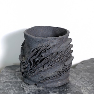 May include: A dark grey ceramic vase with a textured, layered design. The vase has a wide, open top and a rough, organic appearance. The surface has a rippled effect, adding visual interest to this unique piece. The vase is approximately 15 cm tall.