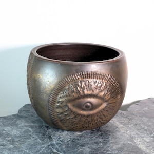 Peut inclure: Un bol en céramique de couleur bronze avec une surface texturée et un grand motif d'œil en relief. Le bol a une forme arrondie et un intérieur sombre. Le motif de l'œil est entouré de lignes rayonnantes.