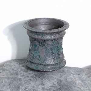 May include: A dark gray, textured vase with a flared base and rim. The vase has a weathered appearance with patches of green, giving it an aged, antique look. The vase is sitting on a gray stone surface.