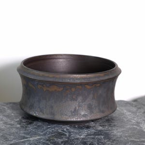 Peut inclure: Un bol en céramique gris foncé, fait à la main, avec un corps large et incurvé et un bord légèrement évasé. Le bol a un éclat métallique avec des reflets bronze et repose sur une surface en pierre grise. L'intérieur est brun foncé.