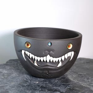 Peut inclure: Un bol en céramique gris foncé avec un motif de visage sculpté. Le bol présente des dents blanches, des yeux orange et jaunes et un bijou bleu. Le bol est posé sur une surface gris foncé.