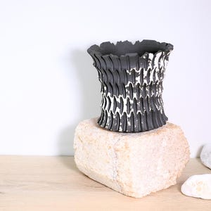 Peut inclure: Un vase en céramique noir et blanc avec un motif texturé et nervuré. Le vase est posé sur une pierre de couleur claire, mettant en valeur sa forme unique et ses couleurs contrastées. Le vase mesure environ 15 cm de haut.