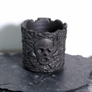 Peut inclure: Un récipient cylindrique noir avec une surface texturée et un motif de crâne. Le crâne a des flammes et des os croisés. Le récipient est fait d'un matériau sombre et repose sur une surface sombre.
