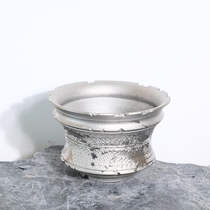 Puede incluir: Un cuenco de metal plateado hecho a mano con un diseño texturizado y martillado alrededor de la base. El cuenco tiene un borde ancho y ligeramente irregular y se apoya sobre una superficie gris texturizada. El diseño sugiere una estética rústica o artesanal.