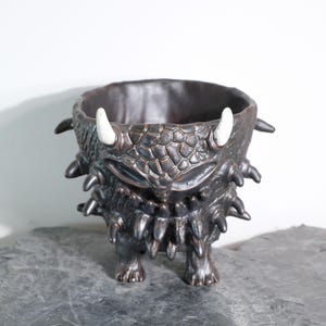 Peut inclure: Un pot de fleurs en céramique de couleur bronze foncé avec un design de monstre. Le pot présente une surface texturée, des cornes blanches et des détails pointus autour du bord et de la base. Il repose sur quatre petits pieds et a une large ouverture.