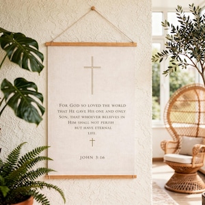 May include: A beige canvas wall hanging with a wooden frame and a gold cross design. The text "FOR GOD SO LOVED THE WORLD...JOHN 3:16" is printed in black. The wall hanging is suspended by a natural twine.