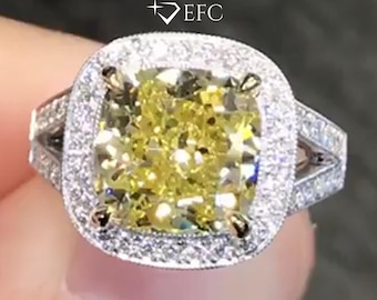 May include: A diamond ring featuring a large, square-cut yellow central stone, encircled by a halo of smaller white diamonds. The band is also embellished with smaller diamonds. The ring is set in a white metal.