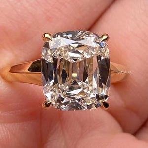 May include: A gold ring featuring a large, cushion-cut diamond. The diamond has a brilliant, multifaceted appearance, reflecting light. The ring's band is a simple, polished gold. The overall design is elegant and classic.