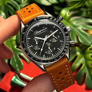 May include: A wristwatch with a black face and a brown leather strap. The watch face features white hands and markers, with the text "Sleepmaster - Professional". The strap has a perforated design.