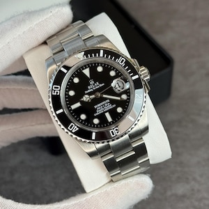 Puede incluir: Un reloj de pulsera de acero inoxidable con esfera negra y bisel giratorio. El reloj presenta una correa plateada y el texto "RELAX Nothing To See Here" en la esfera. El reloj se muestra sobre una superficie blanca.
