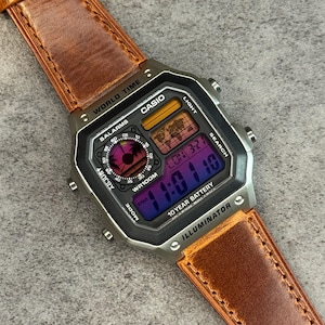 May include: A Casio digital watch with a brown leather strap. The watch face displays multiple digital screens with the time, date, and other functions. The watch has a black case and features the text "WORLD TIME", "CASIO", and "ILLUMINATOR".