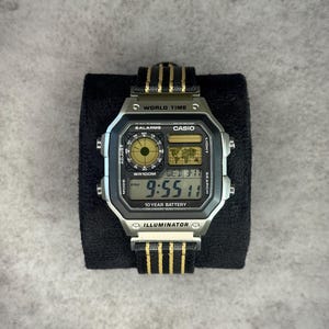 Puede incluir: Un reloj digital Casio plateado con una correa a rayas negras y doradas. La esfera del reloj muestra la hora como 21:55, junto con la fecha y las funciones de hora mundial. El reloj está etiquetado como "ILLUMINATOR" y "10 YEAR BATTERY".