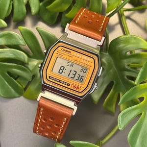Puede incluir: Un reloj digital con caja rectangular plateada y esfera naranja con el texto CASIO, ALARM CHRONO y WATER RESIST. El reloj muestra la hora 8:13:36 y el día SA 30. Tiene una correa de cuero marrón perforada.