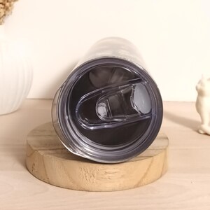 May include: A stainless steel tumbler with a black interior and a clear plastic lid. The lid has a sliding closure and a small opening for drinking. The tumbler is resting on a small, round wooden coaster.