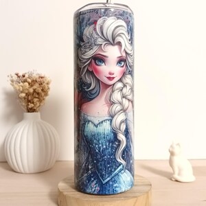 May include: A stainless steel tumbler featuring a detailed illustration of a princess with white hair, blue eyes, and a blue dress. The tumbler has a silver straw and is set on a wooden coaster. The design includes a crown and a braided hairstyle.