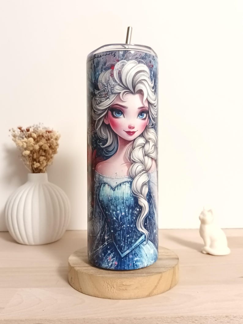 May include: A stainless steel tumbler featuring a detailed illustration of a princess with white hair, blue eyes, and a blue dress. The tumbler has a silver straw and is set on a wooden coaster. The design includes a crown and a braided hairstyle.