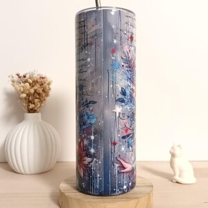 May include: A stainless steel tumbler with a floral and script design. The tumbler features a blue, gray, and white color scheme with red and blue floral accents. A silver straw is inserted into the top of the tumbler. The tumbler is displayed on a wooden coaster.