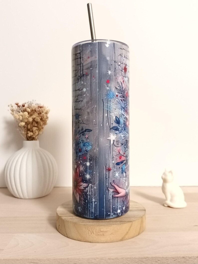 May include: A stainless steel tumbler with a floral and script design. The tumbler features a blue, gray, and white color scheme with red and blue floral accents. A silver straw is inserted into the top of the tumbler. The tumbler is displayed on a wooden coaster.