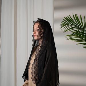 Puede incluir: Un velo negro con intrincados bordes de encaje. El velo está drapeado sobre la cabeza y los hombros, creando un aspecto dramático y elegante. Los detalles de encaje añaden un toque de sofisticación al diseño general. El velo está hecho de una tela suave y fluida.