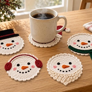 Puede incluir: Un conjunto de cinco posavasos redondos de ganchillo con forma de muñeco de nieve, con accesorios de invierno como un sombrero de copa negro, orejeras rojas, una bufanda verde y un gorro de Papá Noel, sobre caras de color crema con nariz naranja.