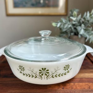 Puede incluir: Fuente blanca para horno con tapa de cristal transparente. El plato presenta un diseño floral verde alrededor del borde. El plato está sobre una tabla de cortar de madera. Una imagen enmarcada y vegetación están en el fondo.
