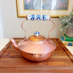 May include: A copper tea kettle with a wooden lid and a white handle decorated with blue floral designs. The kettle sits on a wooden cutting board. The handle is supported by copper arches.