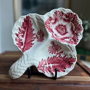 Puede incluir: Una fuente blanca en forma de hoja con asa, con un diseño floral burdeos. La fuente está sostenida por un soporte de metal negro y se encuentra sobre una superficie de madera. El patrón floral se repite en la fuente.