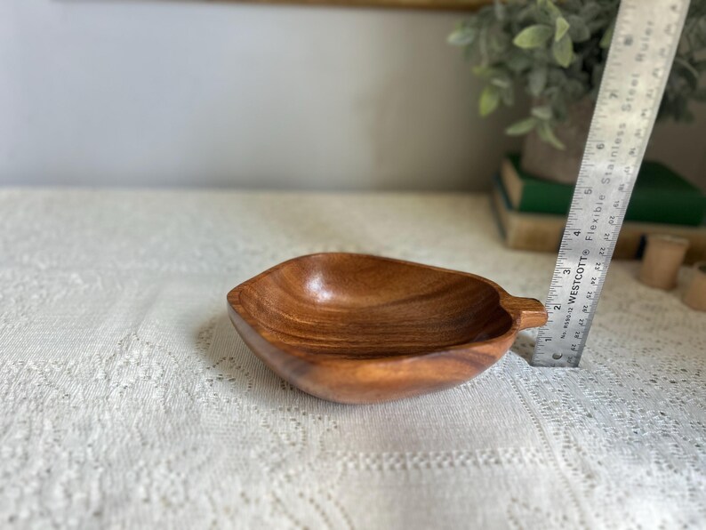 K&ouml;nnte beinhalten: Eine holzfarbene, blattf&ouml;rmige Schale mit Griff. Die Schale besteht aus hellbraunem Holz und hat eine glatte, polierte Oberfl&auml;che. Ein Metalllineal befindet sich neben der Schale. Die Schale ist ca. 18 cm lang.