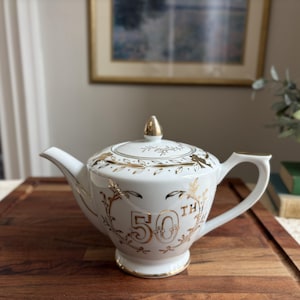 May include: White ceramic teapot with gold accents and a lid. The teapot features the number "50th" in gold script, along with decorative gold floral and bird designs. The teapot is on a wooden surface.