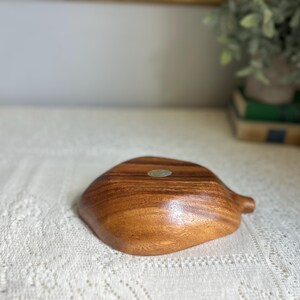 K&ouml;nnte beinhalten: Eine Holzschale mit nat&uuml;rlicher Maserung. Die Schale hat eine organische Form mit einem kleinen griffartigen Ansatz. Ein kleiner, runder Aufkleber ist sichtbar. Die Schale dient wahrscheinlich zum Servieren oder Ausstellen. Das Holz hat einen warmen, braunen Ton.