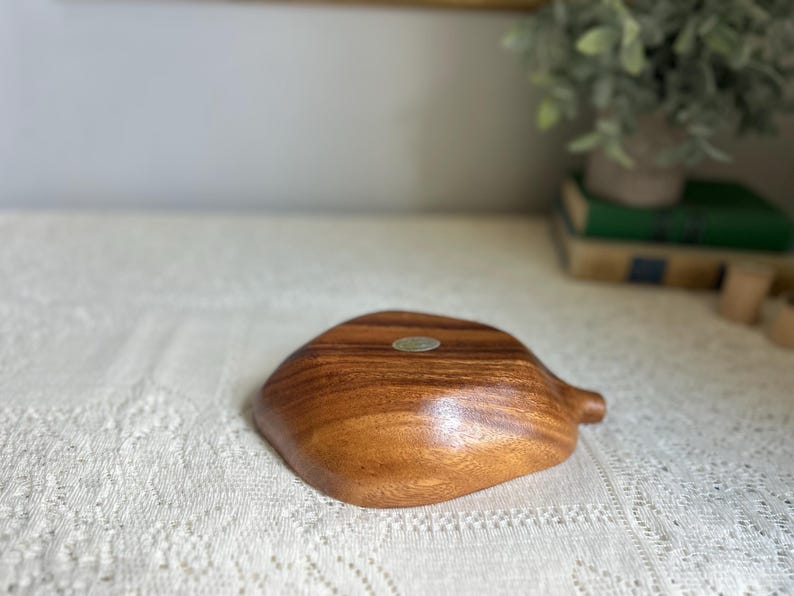 K&ouml;nnte beinhalten: Eine Holzschale mit nat&uuml;rlicher Maserung. Die Schale hat eine organische Form mit einem kleinen griffartigen Ansatz. Ein kleiner, runder Aufkleber ist sichtbar. Die Schale dient wahrscheinlich zum Servieren oder Ausstellen. Das Holz hat einen warmen, braunen Ton.