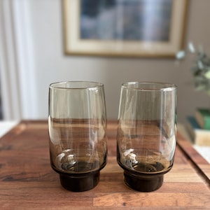May include: Two clear, tinted brown drinking glasses with a dark brown base. The glasses are cylindrical with a slightly flared top. They are displayed on a wooden surface, with a blurred background including a framed picture and greenery.