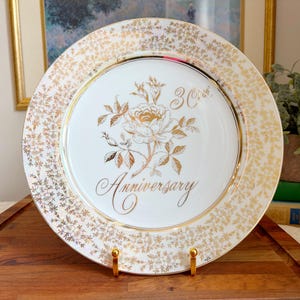 Può includere: Un piatto decorativo bianco e oro con un motivo floreale e la parola "Anniversary" in elegante scrittura. Il piatto ha un bordo dorato e un motivo floreale dorato sul bordo. È visibile anche il numero "30".